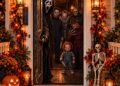 A Halloween front porch at night with costumed horror movie characters standing in an open doorway, glowing jack-o-lanterns, a seated skeleton, fall garlands, and a welcome mat.
