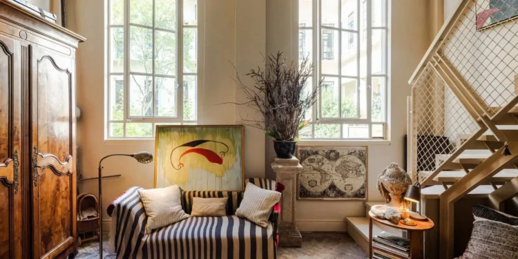 Historic London apartment with tall windows, aged parquet flooring, an antique wooden armoire, striped sofa, modern artwork, and a contemporary metal staircase.